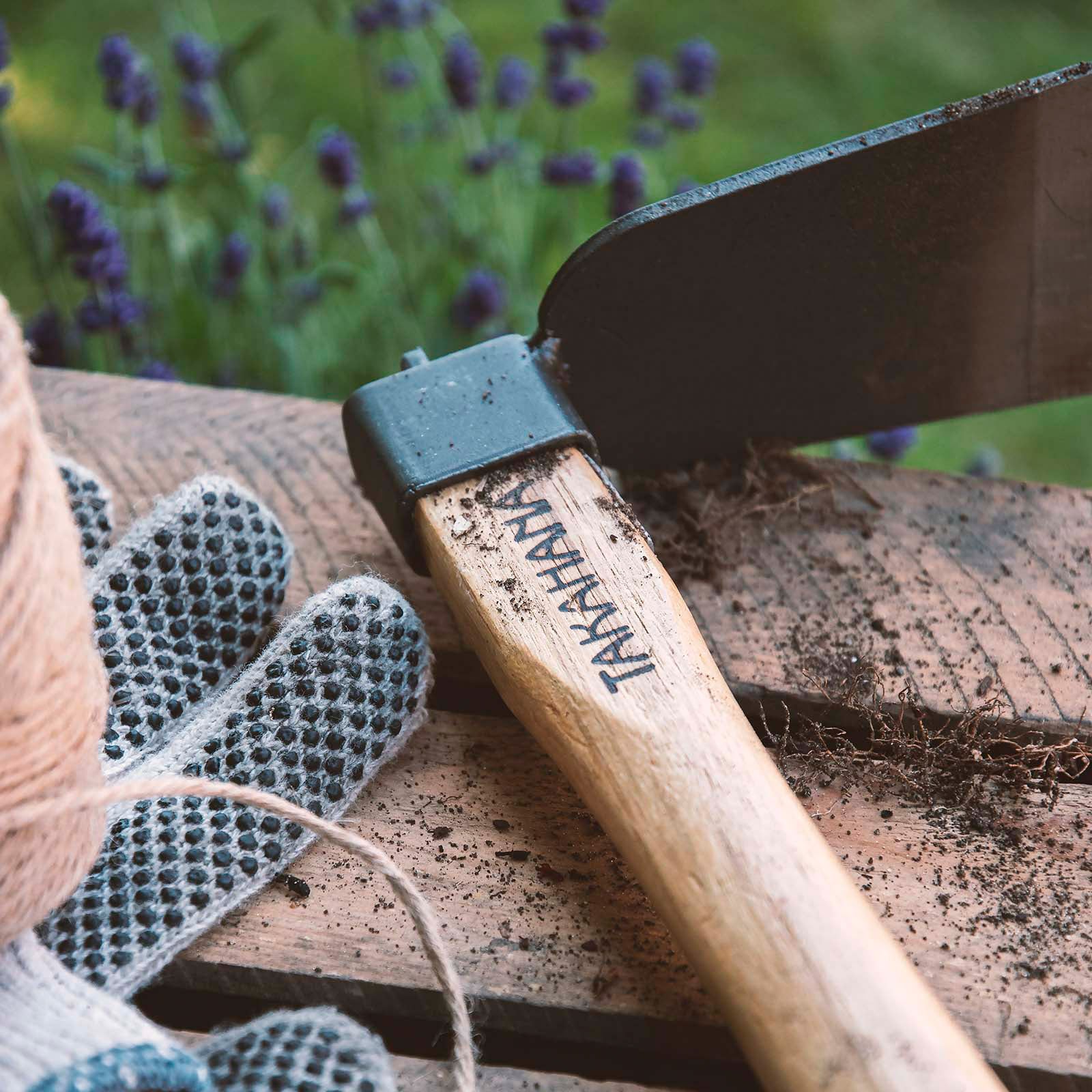 Takahana Pflanzhacke mit etwas Erde bedeckt auf einer Holzkiste abgelegt. Daneben liegen Gartenhandschuhe